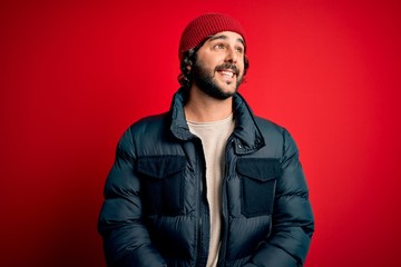 Young handsome man with beard wearing casual coat and wool cap over red background looking away to side with smile on face, natural expression. Laughing confident.