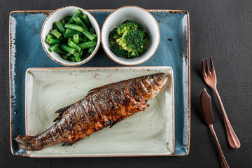 Baked trout fish with spices, green beans, broccoli in plate on black background. Healthy food, dieting, sports nutrition, top view