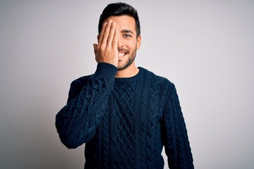 Young handsome man with beard wearing casual sweater standing over white background covering one eye with hand, confident smile on face and surprise emotion.