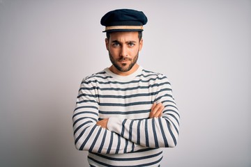 Young handsome sailor man with beard wearing navy striped uniform and captain hat skeptic and nervous, disapproving expression on face with crossed arms. Negative person.