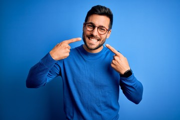 Young handsome man with beard wearing casual sweater and glasses over blue background smiling cheerful showing and pointing with fingers teeth and mouth. Dental health concept.
