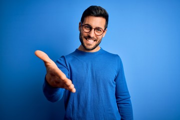 Naklejka premium Young handsome man with beard wearing casual sweater and glasses over blue background smiling cheerful offering palm hand giving assistance and acceptance.