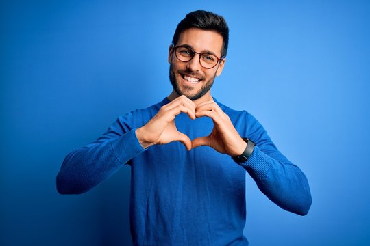 Young handsome man with beard wearing casual sweater and glasses over blue background smiling in love doing heart symbol shape with hands. Romantic concept.