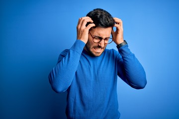 Young handsome man with beard wearing casual sweater and glasses over blue background suffering from headache desperate and stressed because pain and migraine. Hands on head.