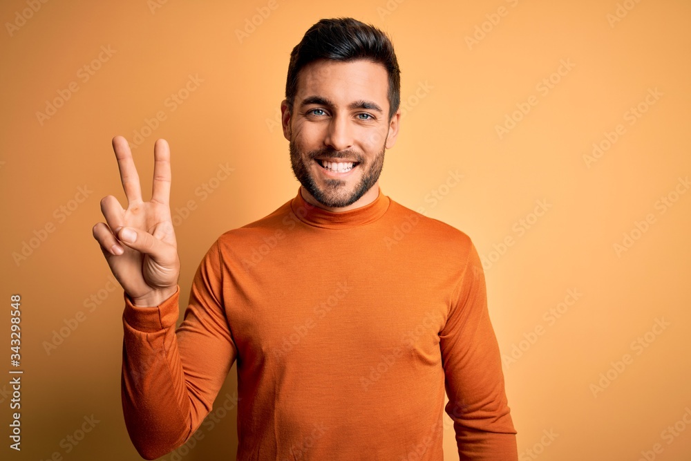 Wall mural Young handsome man with beard wearing casual sweater standing over yellow background showing and pointing up with fingers number two while smiling confident and happy.