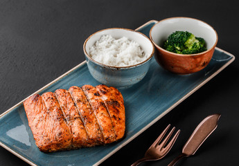 Baked fish fillet with spices, boiled rice and broccoli in plate on black background. Healthy food, dieting, sports nutrition, top view