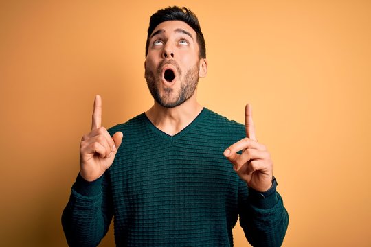 Young handsome man with beard wearing casual sweater standing over yellow background amazed and surprised looking up and pointing with fingers and raised arms.