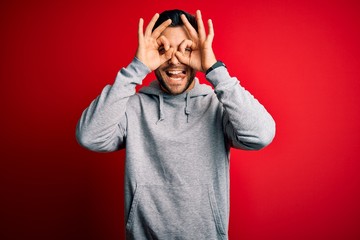Young handsome sportsman wearing sweatshirt standing over isolated red background doing ok gesture like binoculars sticking tongue out, eyes looking through fingers. Crazy expression.