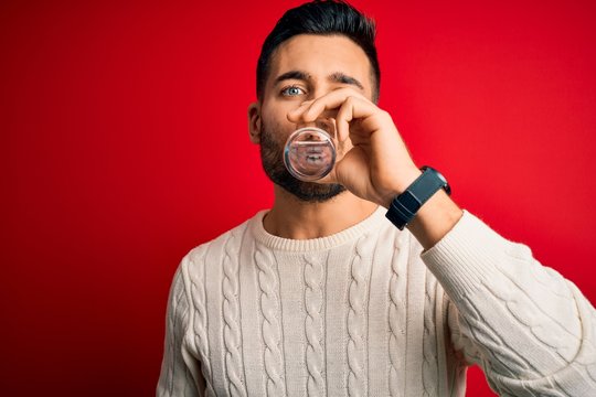 Young handsome man drinking glass of healthy water to refreshment standing over isolated red background