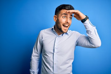 Young handsome man wearing elegant shirt standing over isolated blue background very happy and smiling looking far away with hand over head. Searching concept.