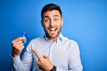 Young handsome man holding blue ribbon as prostate campaing support over blue background very happy...