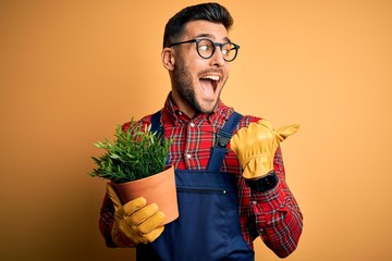 Young gardener man wearing working apron gardening plat for hobby over yellow background pointing...