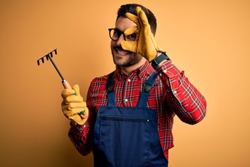 Young gardener man wearing working apron using gloves and tool over yellow background with happy...