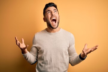 Young handsome man wearing casual sweater standing over isolated yellow background crazy and mad shouting and yelling with aggressive expression and arms raised. Frustration concept.