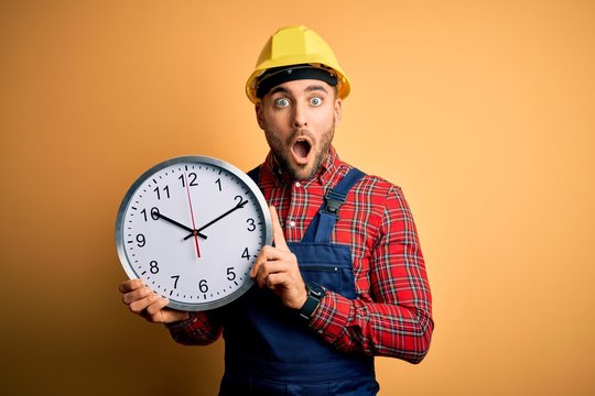Young builder man wearing safety helmet holding big clock over yellow background scared in shock with a surprise face, afraid and excited with fear expression