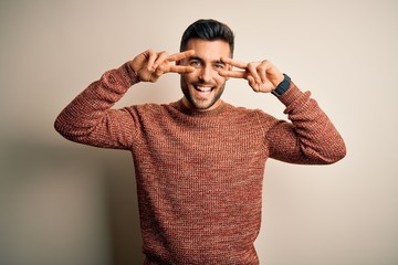 Young handsome man wearing casual sweater standing over isolated white background Doing peace symbol with fingers over face, smiling cheerful showing victory