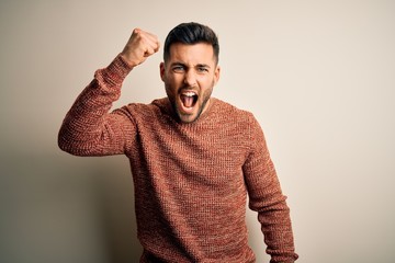 Young handsome man wearing casual sweater standing over isolated white background angry and mad raising fist frustrated and furious while shouting with anger. Rage and aggressive concept.