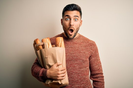 Young handsome man holding groceries paper bag of fresh baguette bread over isolated background scared in shock with a surprise face, afraid and excited with fear expression