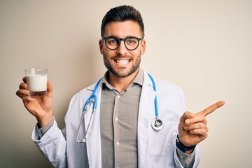 Young doctor man wearing stethoscope holding a glass of milk over isolated background very happy pointing with hand and finger to the side