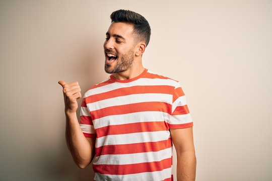 Young handsome man wearing casual striped t-shirt standing over isolated white background smiling with happy face looking and pointing to the side with thumb up.