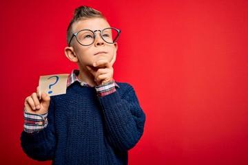Young little caucasian kid wearing glasses holding paper note with question mark sign serious face...