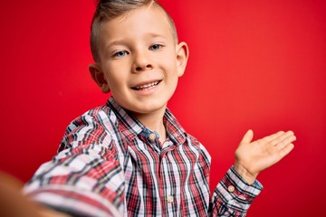 Close up of young little caucasian kid with blue eyes taking a selfie photo over red background very happy and excited, winner expression celebrating victory screaming with big smile and raised hands