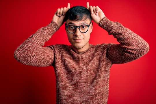 Young handsome hispanic man wearing nerd glasses over red background doing funny gesture with finger over head as bull horns