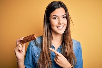 Young beautiful girl holding healthy protein bar standing over isolated yellow background very...