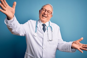 Senior grey haired doctor man wearing stethoscope and medical coat over blue background looking at the camera smiling with open arms for hug. Cheerful expression embracing happiness.