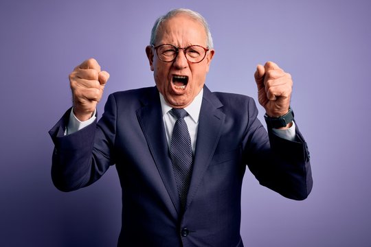 Grey haired senior business man wearing glasses and elegant suit and tie over purple background angry and mad raising fists frustrated and furious while shouting with anger. Rage and aggressive