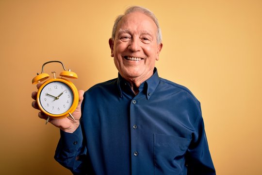 Senior grey haired man holding vintage alarm clock over yellow background with a happy face standing and smiling with a confident smile showing teeth
