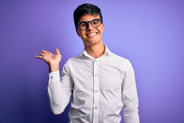 Naklejka premium Young handsome business man wearing shirt and glasses over isolated purple background smiling cheerful presenting and pointing with palm of hand looking at the camera.