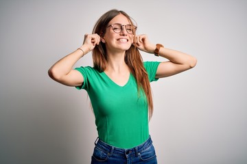 Young beautiful redhead woman wearing casual green t-shirt and glasses over white background Smiling pulling ears with fingers, funny gesture. Audition problem