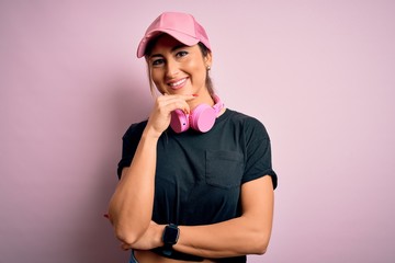 Young beautiful fitness sports woman wearing training cap and headphones over pink background looking confident at the camera smiling with crossed arms and hand raised on chin. Thinking positive.
