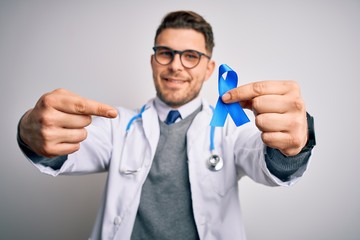 Young doctor man wearing medical coat and holding colon cancer awareness blue ribbon very happy pointing with hand and finger