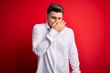 Young business man with blue eyes wearing elegant shirt standing over red isolated background smelling something stinky and disgusting, intolerable smell, holding breath with fingers on the nose. Bad