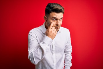 Young business man with blue eyes wearing elegant shirt standing over red isolated background Pointing to the eye watching you gesture, suspicious expression