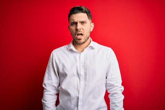 Young business man with blue eyes wearing elegant shirt standing over red isolated background In shock face, looking skeptical and sarcastic, surprised with open mouth