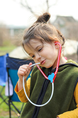 A small, beautiful, cute girl blows into the tube of a toy stethoscope. Dark-haired white girl plays a doctor outdoors