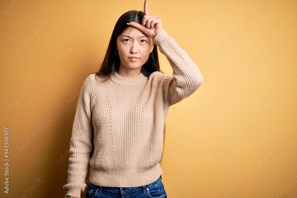 Poster Young beautiful asian woman wearing casual sweater over yellow isolated background making fun of people with fingers on forehead doing loser gesture mocking and insulting.