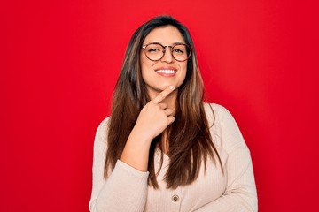 Young hispanic smart woman wearing glasses standing over red isolated background looking confident at the camera with smile with crossed arms and hand raised on chin. Thinking positive.