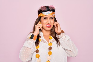 Young hispanic hippie woman wearing fashion boho style and sunglasses over pink background covering ears with fingers with annoyed expression for the noise of loud music. Deaf concept.