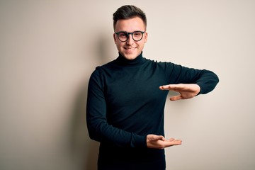 Young handsome caucasian man wearing glasses and casual sweater over isolated background gesturing with hands showing big and large size sign, measure symbol. Smiling looking at the camera. Measuring