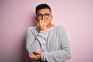 Young handsome latin man wearing casual sweater and glasses over pink background looking stressed and nervous with hands on mouth biting nails. Anxiety problem.