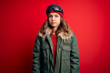 Young blonde girl wearing ski glasses and winter coat for ski weather over red background Relaxed with serious expression on face. Simple and natural looking at the camera.