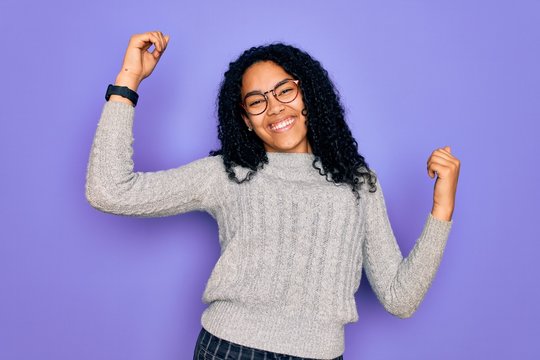 Young african american woman wearing casual sweater and glasses over purple background Dancing happy and cheerful, smiling moving casual and confident listening to music