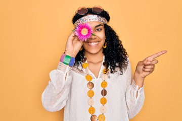 Young african american curly hippie woman wearing sunglasses holding pink flower on eye very happy pointing with hand and finger to the side