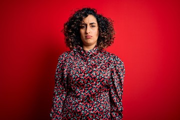 Young beautiful curly arab woman wearing casual floral dress standing over red background skeptic and nervous, frowning upset because of problem. Negative person.