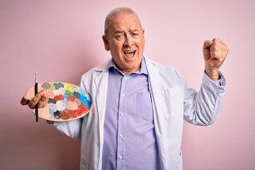 Middle age artist man painting using paintbrush and palette with colors over pink background screaming proud and celebrating victory and success very excited, cheering emotion