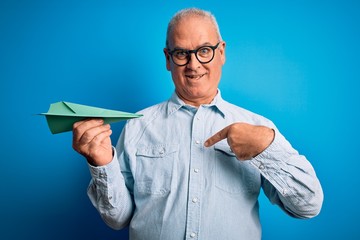 Middle age handsome hoary man holding paper airplane over isolated blue background with surprise...